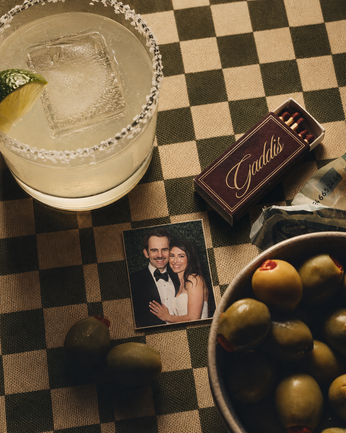 A cocktail, olives, a Gaddis matchbook, and a photograph of Victoria and Sam